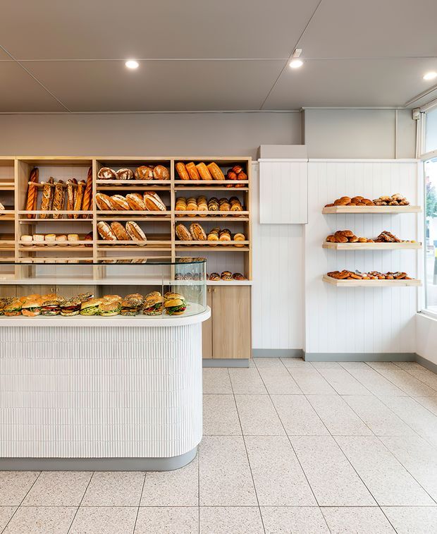 A bright, contemporary bakery or café interior with a curved, vertically fluted white service counter and glass display case in the foreground. Behind it are open timber shelves stocked with bread and pastries, along with wall-mounted white display shelves. The space has pale grey walls, a tiled floor, recessed ceiling lights, and large windows providing natural light.