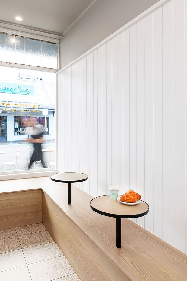 Bright, compact café seating area with a long built-in light timber bench, two small round black pedestal tables, and white vertical wall panelling. A large street-facing window overlooks a neighbouring shop, while terrazzo-style tiled flooring and a simple recessed ceiling light complete the clean fit-out.