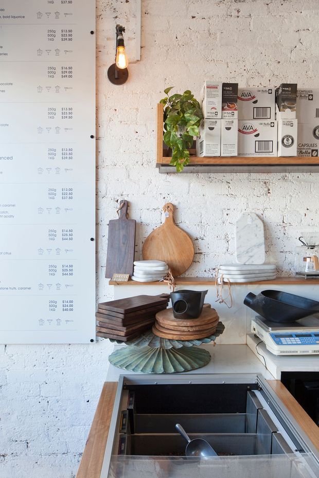 A compact café or takeaway food-service counter with a white-painted exposed brick wall. A large menu board occupies the left side, while open timber shelving displays coffee capsule boxes and supplies. Timber and stone serving boards, ceramic plates, bowls, cups, and a black jug are arranged above a stainless-steel refrigerated or storage counter. A simple exposed-bulb wall light and trailing plant add warmth to the utilitarian workspace.