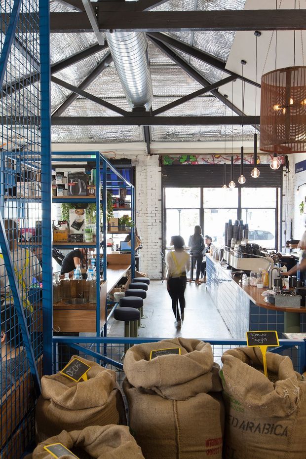 Interior view of a bright industrial-style coffee roastery and café. The space features exposed dark steel roof trusses, translucent insulated roofing, a large exposed silver ventilation duct, white-painted brick walls, blue metal mesh shelving and partitions, a tiled service counter, pendant lights, café stools, coffee equipment, and burlap coffee sacks in the foreground. Large glazed doors at the rear bring in natural light.