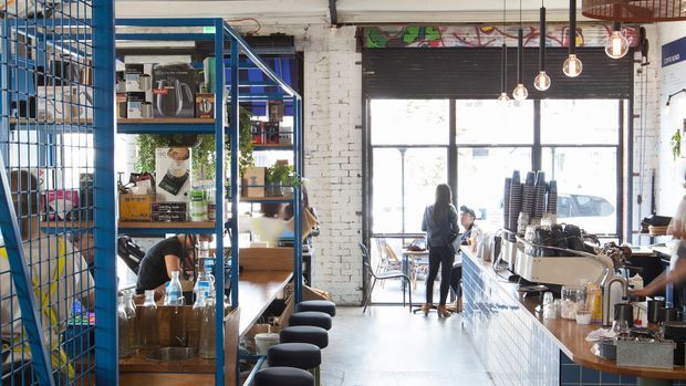Bright industrial-style café interior with a long tiled service counter on the right, exposed white-painted brick walls, large black-framed windows, suspended pendant lights, and open blue metal shelving dividing the seating area. The space includes stools, display shelving, café equipment, and a visible street-facing frontage.