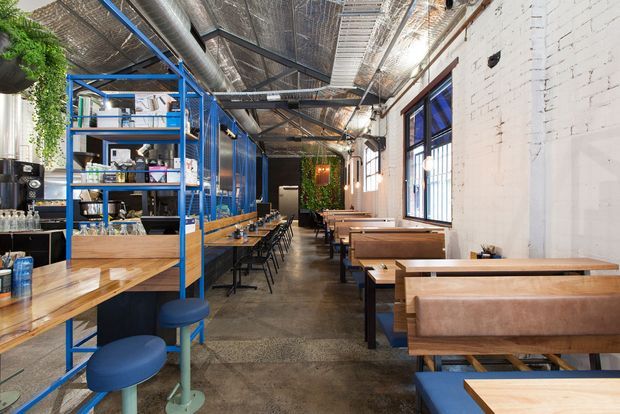 Interior view of an industrial-style restaurant or café with exposed services and ductwork, a polished concrete floor, white-painted brick walls, blue-painted steel shelving and framing, timber tables, and booth seating. The long narrow dining space includes a visible bar or service area, abundant greenery, and large windows.