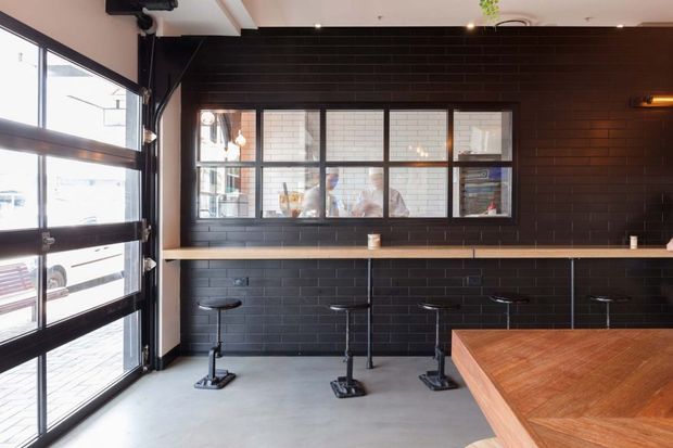 A contemporary commercial bar or café interior with a dark tiled feature wall, long timber counter, black bar stools, large industrial-style garage glazing, and an interior service window. The space has a raw, streamlined fit-out with exposed structural and mechanical elements.