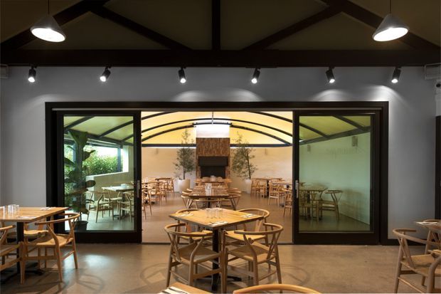 A warmly lit contemporary restaurant dining area viewed from the main room toward a glazed private or adjoining dining space. Timber tables and chairs fill the foreground, while dark-framed full-height glass partitions, track lighting, exposed dark ceiling beams, and a central fireplace or feature wall define the interior. The palette is restrained with grey walls, timber furniture, and warm amber lighting.