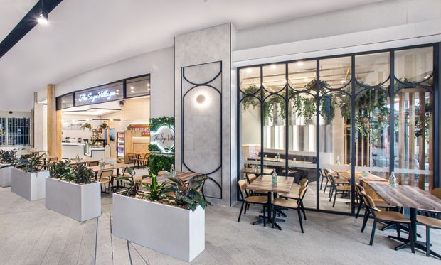 A bright contemporary indoor commercial dining and retail environment, with café tables and chairs arranged beside a glazed storefront. Large white rectangular planters with leafy plants divide the seating area, while black-framed glass partitions, timber details, decorative metalwork, and a feature wall light add visual interest. A food or beverage shop is visible beyond the open storefront.