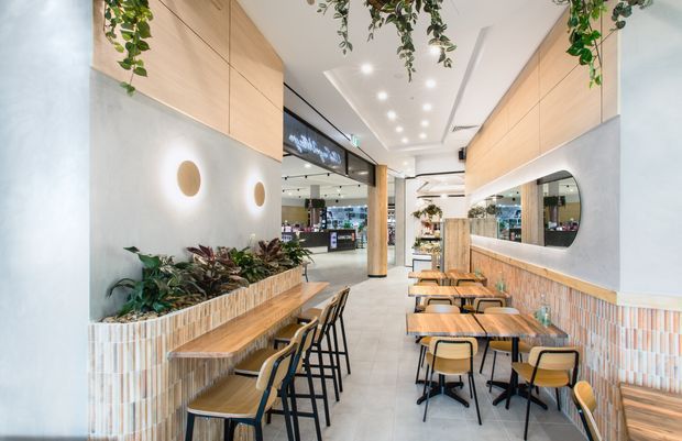 Bright contemporary café interior with a narrow dining layout, pale timber wall cabinetry, integrated banquette seating, small timber tables, black metal-framed chairs, curved tiled planter and bench fronts, large rounded mirrors, and hanging greenery. The space opens toward a shopping or commercial area in the background.