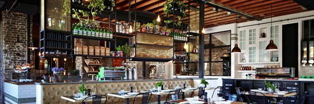 A stylish industrial restaurant or café interior with exposed brick walls, a dark timber slatted ceiling, black metal-framed shelving, pendant lighting, a long tufted banquette, and a visible bar and service counter. Tables and chairs occupy the foreground, while greenery and glassware decorate the open shelving.