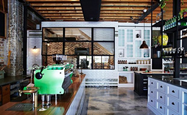A stylish industrial café or commercial hospitality interior with a green espresso machine on a timber counter, exposed timber ceiling beams, polished concrete flooring, exposed brick walls, black-framed glazed partitions, white subway tile, open black shelving, and pale blue built-in cabinetry.