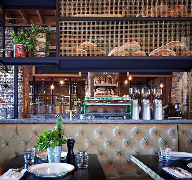 A stylish restaurant or café interior viewed from the dining area, featuring a tufted leather banquette, dark tables set with plates and glasses, and a bar counter with a coffee machine and display shelves of bread. Behind the counter are exposed brick and timber details, pendant lighting, dark metal framing, and a wire-mesh bread display. The atmosphere is warm, industrial, and refined.