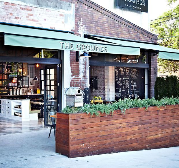 Street-facing café storefront with a weathered red-brick facade, dark-framed glazing and doors, green awnings, and a large built-in timber planter box filled with greenery. The open frontage reveals a bar and seating area inside, with chalkboard-style menu graphics and warm pendant lighting.
