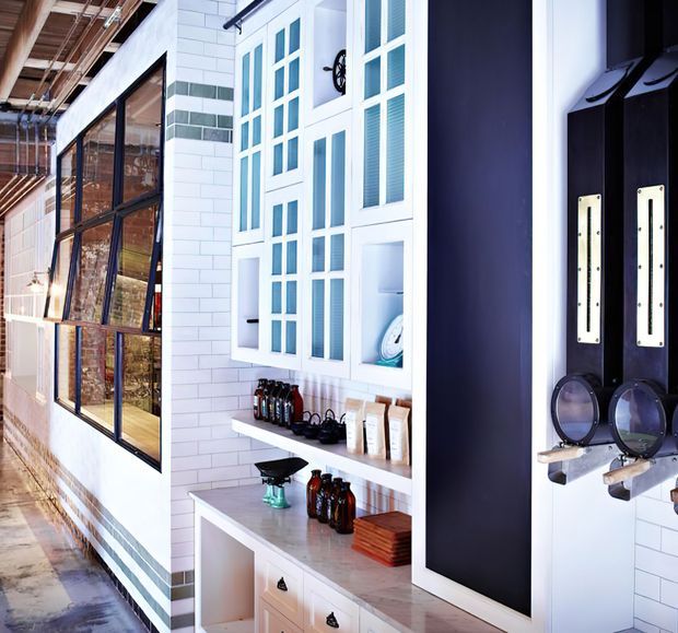 Bright, eclectic commercial-style interior with white subway-tiled walls, dark-framed glazing, and a built-in white display and storage cabinet. Open shelving holds bottles and small retail or grooming items, while a large dark vertical panel and black wall-mounted fixtures add contrast. The exposed ceiling structure and industrial glazing suggest a converted warehouse or industrial-inspired fit-out.