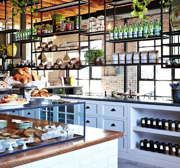 A bright industrial-style bakery or café interior with a white tiled service counter, timber benchtops, pale blue-grey cabinetry, exposed brick walls, black metal-framed windows, and suspended black metal shelving displaying bread, bottled drinks, jars, and packaged goods. A glass pastry display case occupies the foreground.