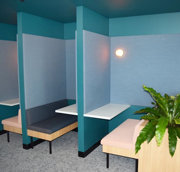A compact contemporary commercial-style breakout or work area with teal partition walls and columns, pale blue acoustic wall panels, dark upholstered bench seating, white work surfaces, light timber bases, and grey carpet. A warm circular wall light and large leafy plant add visual accents. The booth-like arrangement suggests a flexible office or collaborative workspace.