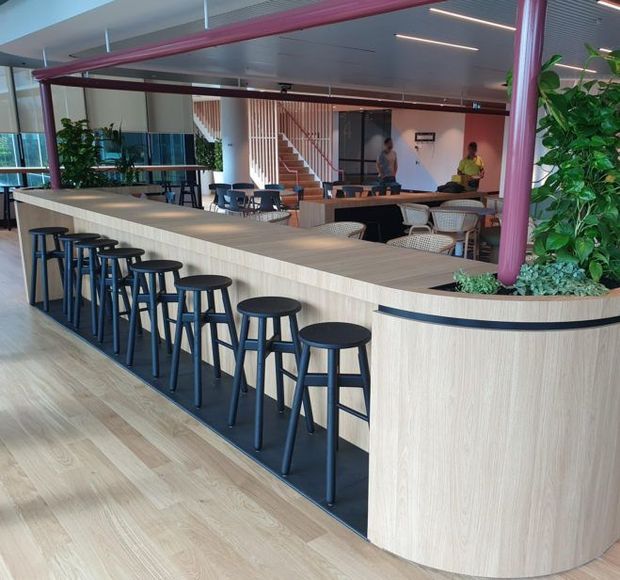 Contemporary commercial hospitality interior featuring a long curved light-wood communal counter with integrated planting, dark bar stools, café tables and chairs, exposed pink-painted structural beams and columns, timber flooring, and a stairway in the background. The open-plan space is bright, refined, and biophilic in character.