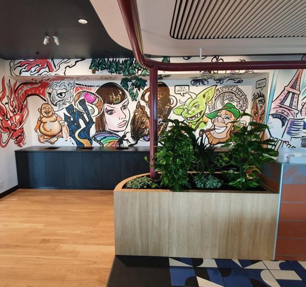 A colourful commercial interior lobby or reception area featuring a large illustrated graffiti-style mural, dark built-in cabinetry, timber flooring, a substantial timber planter with lush indoor plants, and exposed red-painted pipework. The ceiling combines dark painted surfaces with linear slatted detailing, while geometric patterned tiles appear in the foreground.