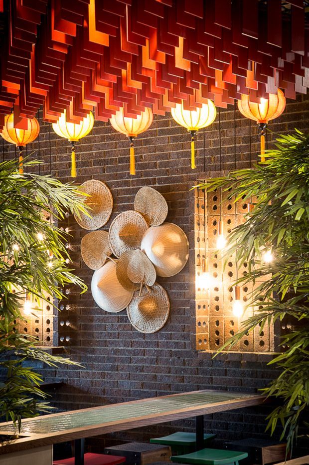 A richly decorated dark brick interior with a dramatic red geometric suspended ceiling installation, glowing lantern-style pendant lights, bamboo-like greenery, woven circular wall art, and a long communal table with colourful stools. The space has an atmospheric, eclectic hospitality or dining feel.