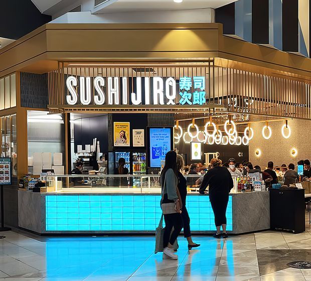 A brightly lit sushi restaurant kiosk in a shopping centre, featuring a prominent illuminated “SUSHI IRO” sign with Japanese characters, warm timber screening and framing, a marble-look service counter, glass food display, turquoise illuminated counter panels, and decorative circular pendant lights. Customers and staff occupy the dining and service area.