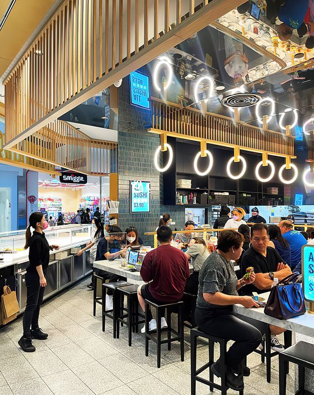 A busy contemporary food-court sushi restaurant interior in a shopping-centre setting. The space features long communal counters, black stools, dark tiled walls, pale stone or solid-surface counters, abundant circular pendant lighting, and extensive timber vertical screening and balustrades around an upper-level circulation or mezzanine area. Diners and staff occupy the foreground and background, with visible sushi and retail signage.