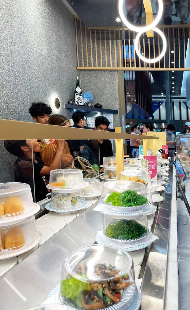 A busy contemporary restaurant or café interior viewed along a stainless-steel conveyor-style food display counter. Covered plates of food dominate the foreground, with diners seated behind a glass and gold-toned divider. The space features textured grey walls, dark shelving, timber-toned vertical balustrade screening, reflective glazing, and distinctive circular ring pendant lights.