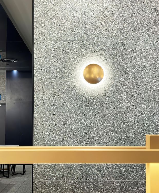 Close-up interior view of a contemporary commercial or hospitality space, dominated by a dark grey speckled stone or terrazzo wall with horizontal panel joints and a glowing spherical brass/gold wall light. A mustard-gold architectural railing or counter edge crosses the foreground, with dark cabinetry or wall panels and part of a tiled floor visible to the left.