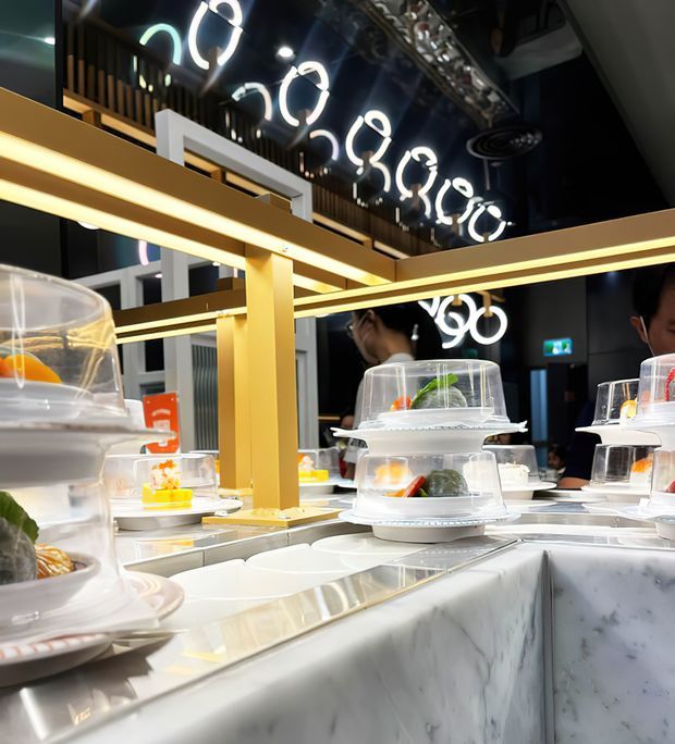 A stylish food-service interior featuring a self-service display counter with plated dishes beneath clear domes. The counter has a white marble-look finish and illuminated gold-toned structural framing. Glossy dark walls and ceiling reflect distinctive circular pendant lights, while staff and additional food displays are visible in the background.