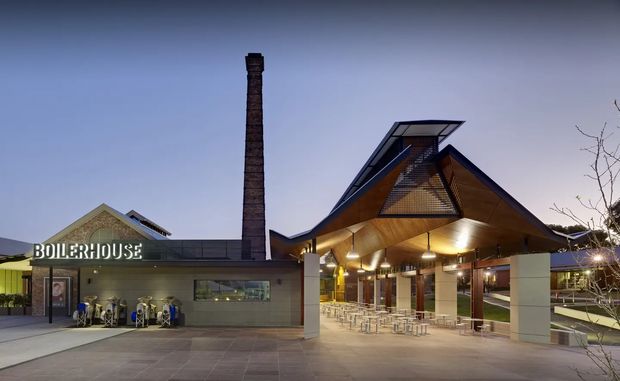 Exterior architectural photograph at dusk showing a contemporary public or commercial building complex. A low flat-roofed volume labelled “BOILERHOUSE” sits beside a tall brick industrial chimney, while the main entrance or covered pavilion has a dramatic steeply pitched, angular roof with exposed timber-toned soffits, clerestory-style glazing, and extensive outdoor seating beneath it. Paved forecourt and surrounding landscaping are visible.