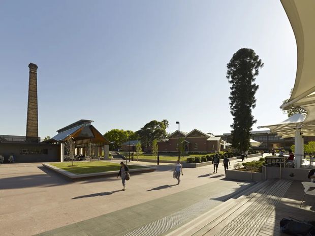 A wide outdoor view of a landscaped public plaza or commercial precinct. Paved pedestrian spaces, low planters, outdoor seating and shade structures frame a cluster of low-rise buildings. A tall brick chimney and contemporary angular entrance canopy are prominent architectural features, with people walking through the open plaza.