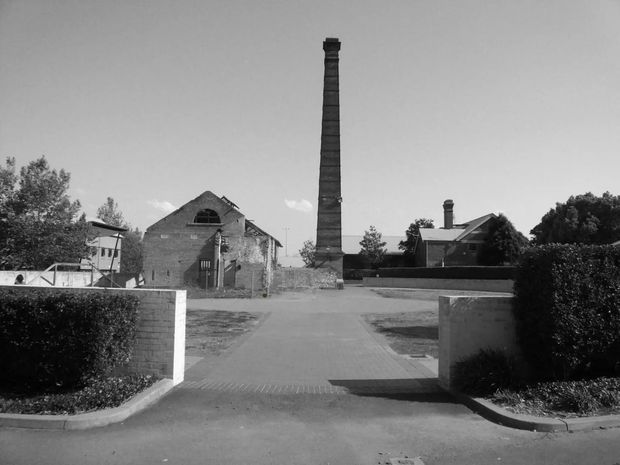 Black-and-white photograph of a historic industrial site, centered on a tall narrow brick chimney beside an old brick building. A paved driveway enters through low brick walls and clipped hedges, with residential or institutional buildings visible behind the site.