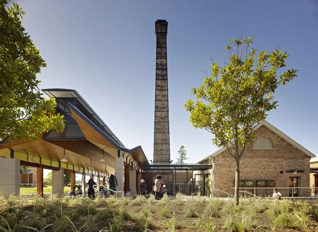 Exterior view of a contemporary adaptive-reuse-style complex, featuring a tall retained brick industrial chimney, a gabled brick building, and a dramatic angular covered pavilion with timber soffits and dark metal roofing. Landscaped planting and people occupy the foreground and courtyard-like space, while glazed openings connect the buildings.