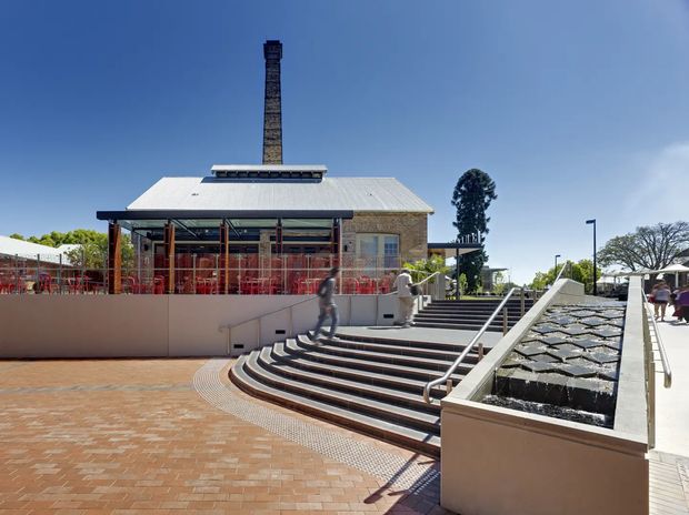 Exterior view of a renovated low-rise brick building with a pitched corrugated roof and prominent tall chimney. The building opens onto a raised terrace with red-framed glazing and outdoor seating, approached by broad curved stairs beside a stepped linear water feature. The surrounding paved plaza and clear blue sky create a bright civic or commercial setting.