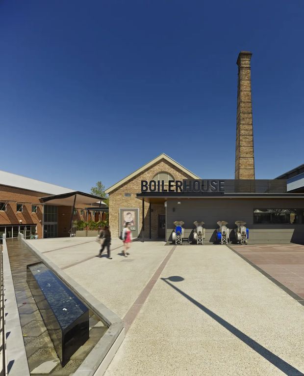 Exterior view of a renovated brick industrial-style boilerhouse building in a public plaza. The facade features a prominent tall masonry chimney, pitched gable roof, large BOILERHOUSE signage, covered service counter, outdoor café-style seating, and a narrow linear water feature in the foreground. The surrounding architecture and paved courtyard suggest an adaptive reuse or commercial hospitality setting.