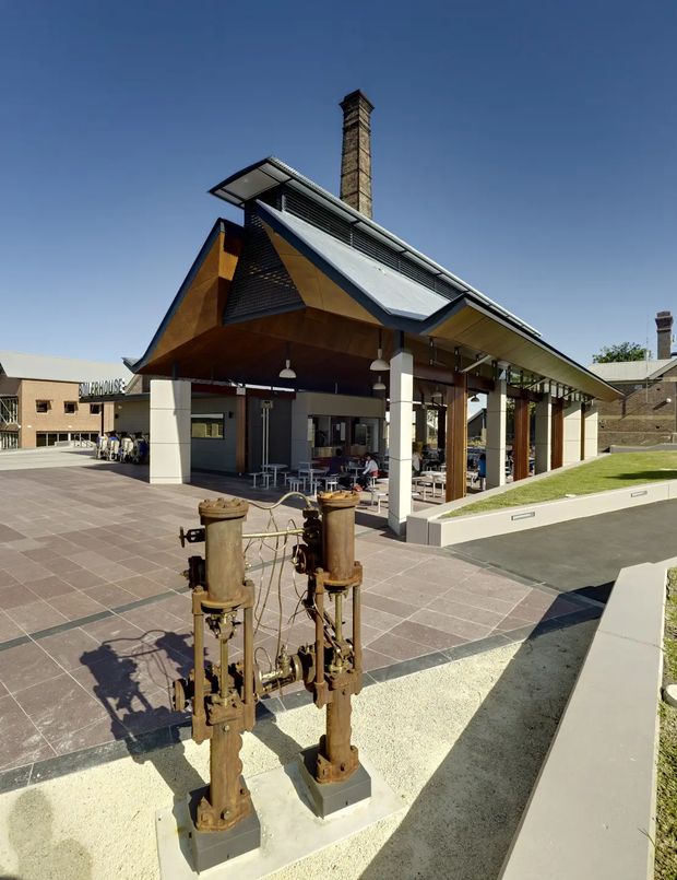 Exterior view of a contemporary pavilion-like commercial building with a dramatic pitched roof, broad covered outdoor seating area, exposed timber soffits, white structural columns, and a tall brick chimney. Historic industrial machinery is displayed in the paved forecourt, with older brick buildings visible in the background.