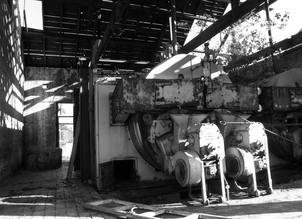 A black-and-white photograph of an industrial mechanical installation in a partially open, weathered structure. Large machinery, exposed beams, rough masonry walls, overhead pipework, and strong sunlight and shadow dominate the scene.
