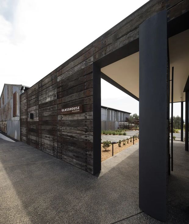 Exterior view of a contemporary commercial-style building entrance clad in dark, weathered timber boards. Large charcoal steel-framed openings and structural columns create a covered passage into a landscaped courtyard, with additional weathered metal-clad structures visible beyond. A small sign reads “GLASSHOUSE RESTAURANT.”