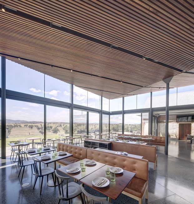 A spacious contemporary dining and hospitality interior with extensive floor-to-ceiling glazing overlooking a rural landscape. Rows of timber dining tables, upholstered banquettes, and chairs fill the room, while a long timber service counter sits beneath a dramatic slatted timber ceiling. Dark slender structural columns, polished concrete or terrazzo flooring, and exposed brickwork at the rear create a refined yet warm restaurant setting.