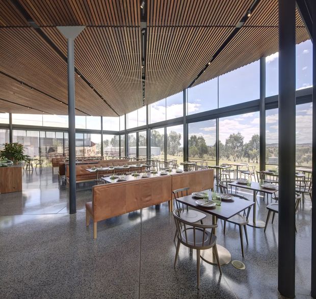 A spacious contemporary dining and hospitality interior with extensive floor-to-ceiling glazing overlooking a landscaped rural setting. The room features a slatted timber ceiling with integrated linear lighting, dark slender structural columns and window frames, polished terrazzo flooring, and a mix of timber dining tables, leather-upholstered banquette seating, and curved timber chairs. The open, airy layout is arranged like a restaurant or café, with communal dining tables and planted built-in areas.
