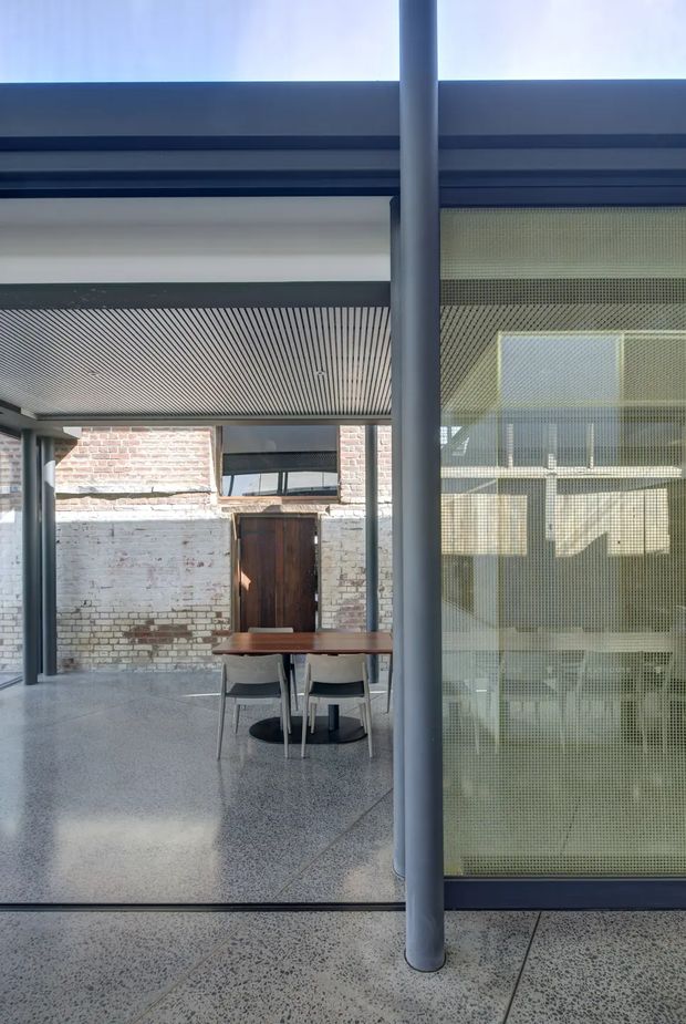 Interior architectural view of a contemporary commercial or institutional space, looking across a polished terrazzo floor toward a dining or meeting table. The room combines exposed and painted brick walls, slender grey structural columns, dark metal framing, a linear slatted ceiling, and a perforated or mesh glazed screen enclosing an adjacent area. Strong daylight enters through high-level glazing, creating a clean, industrial-modern atmosphere.