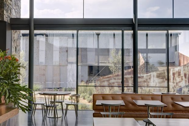 A contemporary café or hospitality interior viewed from inside through expansive floor-to-ceiling glazing. The space contains a long caramel-brown button-tufted banquette, small round café tables with slim dark metal chairs, polished concrete flooring, exposed structural columns, and a large leafy plant. An industrial-style corrugated metal building and courtyard are visible outside through the windows.