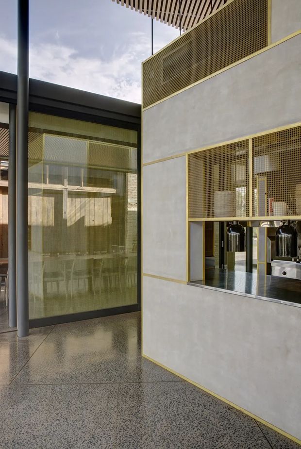 A modern commercial interior courtyard or hospitality space with a polished terrazzo floor, exposed concrete walls, black-framed glazing, and brass-toned perforated metal screens. A service counter with stainless-steel equipment is visible through a large screened opening. Slatted timber ceiling detailing and dark structural columns add visual interest, while the open upper area brings in daylight.