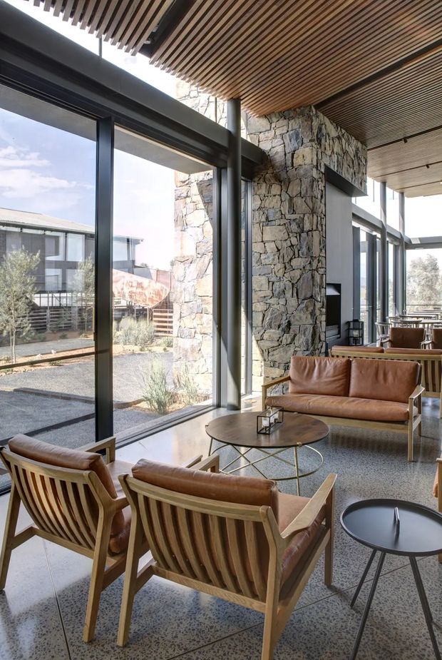 A bright, sophisticated living and lounge area with full-height black-framed glazing, a rugged natural stone feature wall, and a slatted timber ceiling. Leather lounge chairs and a sofa surround round coffee tables, while the polished terrazzo floor and open connection to an outdoor courtyard create an airy indoor-outdoor atmosphere.