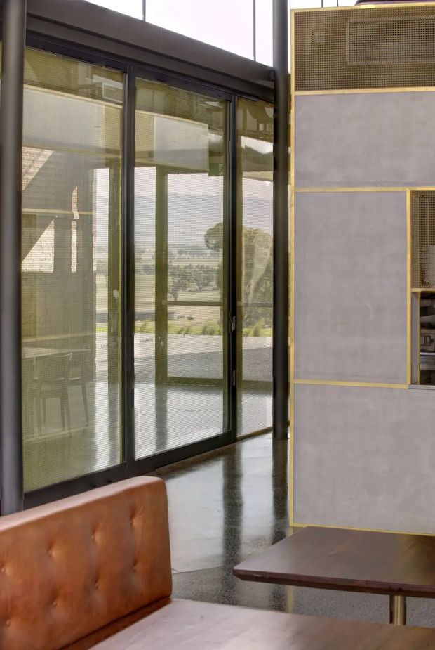 Interior view of a refined modern space with full-height black-framed glazed doors opening toward an outdoor terrace or garden. A polished concrete floor, muted grey feature partition with fine brass-toned trim and perforated screening, and a caramel leather banquette with dark timber table create a sophisticated, lightly industrial atmosphere. Exterior landscape is visible through the glazing.