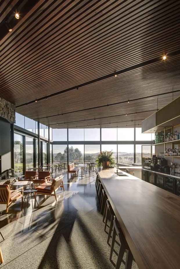 A spacious, sunlit contemporary café or hospitality interior with floor-to-ceiling glazing overlooking a landscaped valley. The room features a long timber-look bar counter with stools, open bottle shelving and coffee equipment, scattered timber-and-leather lounge seating and small tables, polished concrete flooring, and a dark linear slatted ceiling with integrated track and pendant lighting. Exposed stonework is visible at the left edge, while strong afternoon light creates long shadows across the floor.