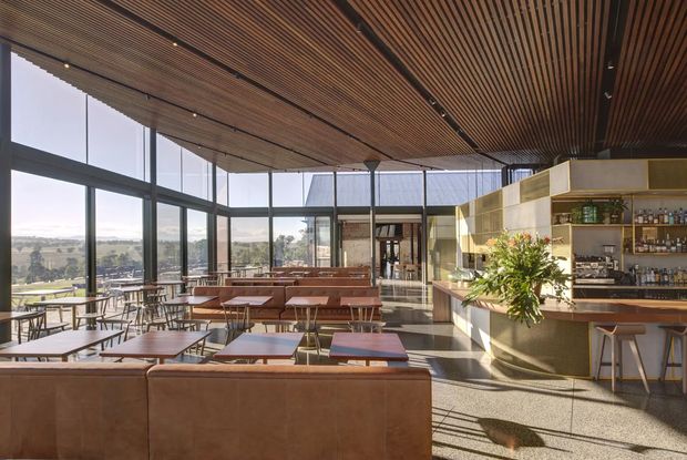 A spacious, sunlit commercial hospitality interior with floor-to-ceiling glazing, expansive countryside views, timber slatted ceiling, polished concrete flooring, leather banquette seating, dining tables, and a built-in bar with open shelving and brass-toned cabinetry. The space has a refined contemporary design with warm natural materials and strong indoor-outdoor visual connection.