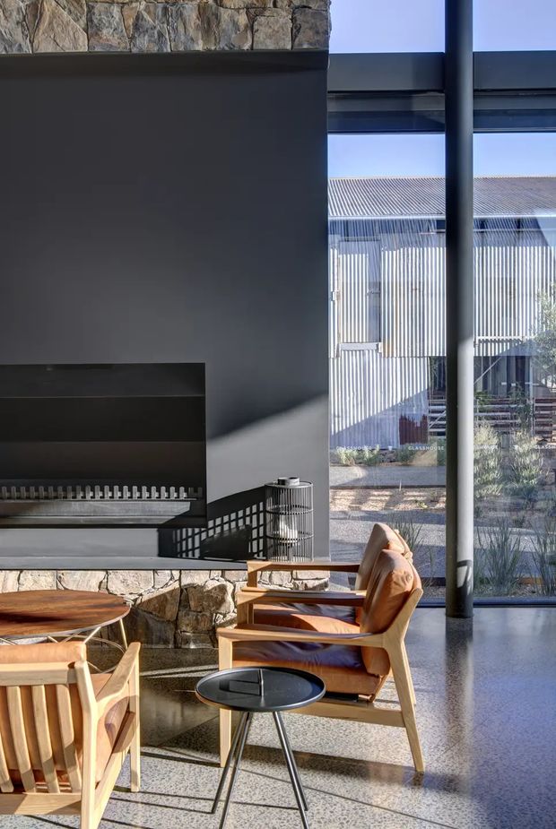 A refined contemporary living room with a dark plaster fireplace wall and substantial rough natural-stone cladding. Timber-framed leather lounge chairs and a low round side table sit on a polished concrete floor. Floor-to-ceiling glazing with dark metal framing opens views to a screened outdoor courtyard or covered structure, bringing strong daylight and geometric shadows into the room.