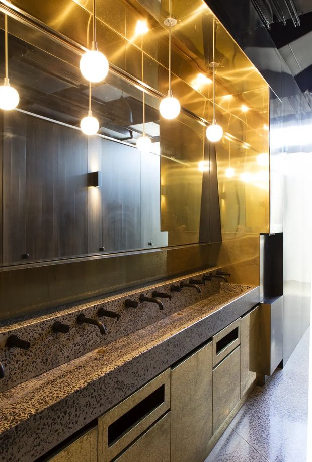 A dramatic contemporary bathroom vanity with a long double-sided or multi-station basin counter, numerous matte-black wall-mounted faucets, and a heavily patterned dark stone or terrazzo-like benchtop and backsplash. Large bronze-gold mirrored panels cover the wall, reflecting suspended spherical pendant lights and dark timber or metal wall finishes. Integrated drawers and cabinetry run beneath the counter, with a bright polished floor and a high-contrast, luxurious commercial-style interior.