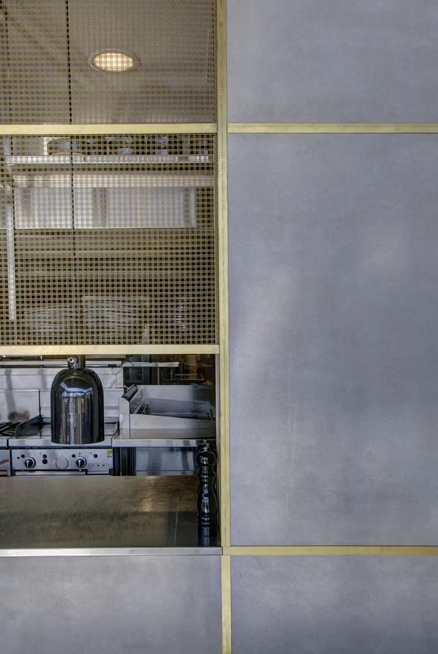 Close-up view of a sophisticated commercial-style kitchen or bar servery. A stainless-steel cooking and preparation area is partially concealed behind a fine brass-toned metal mesh screen, while large grey panelled surfaces with brass trim form the surrounding wall. The image has a refined industrial character with strong geometric lines and muted metallic tones.