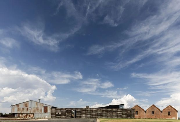Wide exterior view of a low, elongated industrial-style compound. Weathered corrugated metal structures and dark timber or metal fencing span the foreground, with a row of small brick gabled buildings behind. The scene is dominated by an expansive blue sky with scattered clouds.