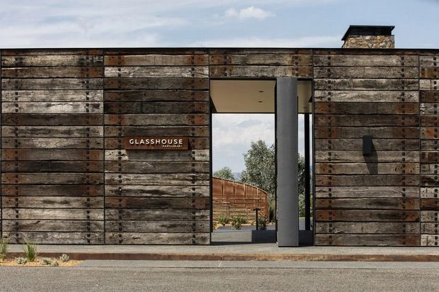 Front exterior view of a contemporary single-storey building with a flat roof and a strongly textured facade clad in reclaimed, weathered timber boards. A tall recessed entry opening is framed by a dark vertical structural element and reveals a landscaped courtyard beyond. The facade includes a small projecting sign reading “GLASSHOUSE” and a dark wall-mounted light.