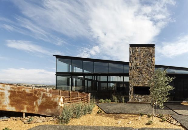 A low, contemporary two-storey home set in a dry, open landscape. The building has extensive dark-framed floor-to-ceiling glazing, a strong horizontal flat roofline, and a prominent rough natural-stone chimney or feature wall with a rectangular fireplace opening. Rusted steel retaining or boundary panels, gravel paving, sparse grasses, and a small olive-like tree create a restrained arid-landscape setting.