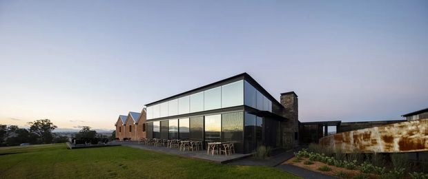 Exterior architectural photograph of a contemporary, multi-part building set on a grassy site. The main structure has a crisp angular roofline, extensive floor-to-ceiling glazing, dark metal framing, and a dark masonry or stone feature wall with chimney. A brick gabled building sits behind it, while a weathered rust-toned metal wall extends to the right. Outdoor tables and chairs line the glazed facade, suggesting a hospitality or commercial use. The scene is photographed in soft early-evening light with a distant landscape visible beyond.